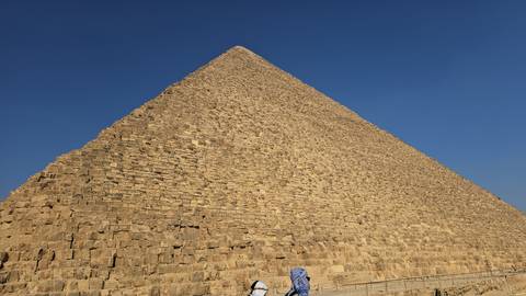       The Great Pyramid of Giza rises steeply into a clear blue sky with small visitors at the base.
  