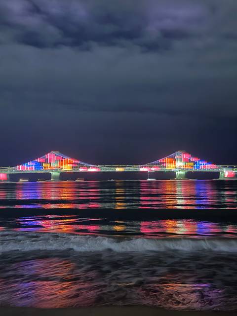       Multicoloured lights illuminate Gwangan Bridge reflecting off calm sea at night.
  