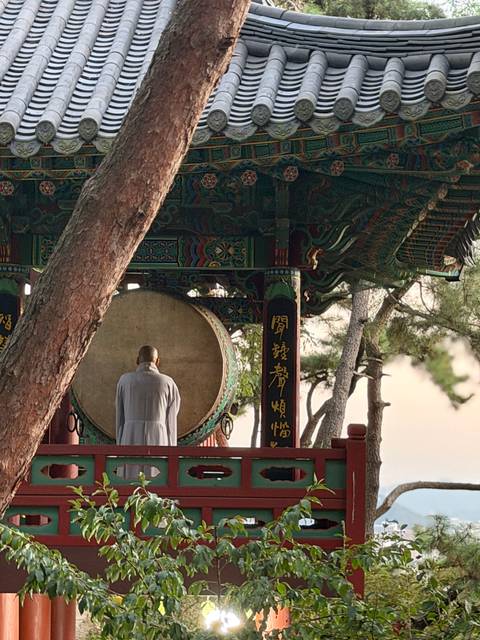       Buddhist monk stands before a giant temple drum inside an ornate pavilion among pines.
  