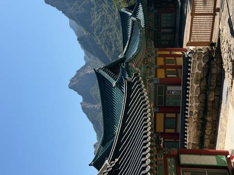       Traditional temple buildings with curved roofs set against jagged mountain peaks and clear sky.
  