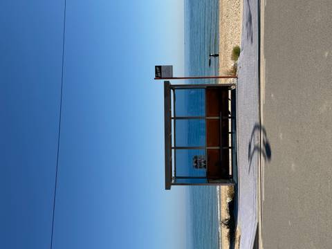       Minimalist coastal bus stop facing a calm blue sea under a clear sky.
  