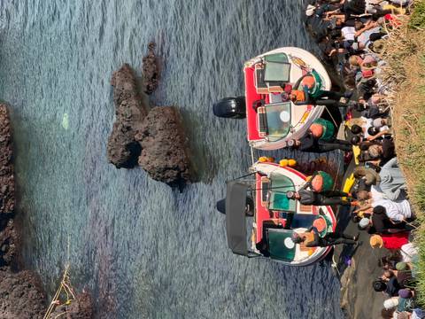       Crowd watching haenyeo divers beside two motorboats docked along rocky Jeju shoreline.
  