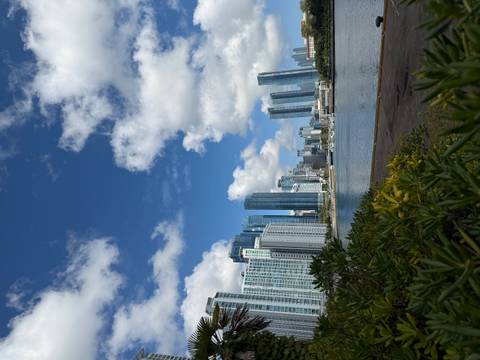       Modern high-rise skyline reflecting in harbor water below scattered clouds.
  