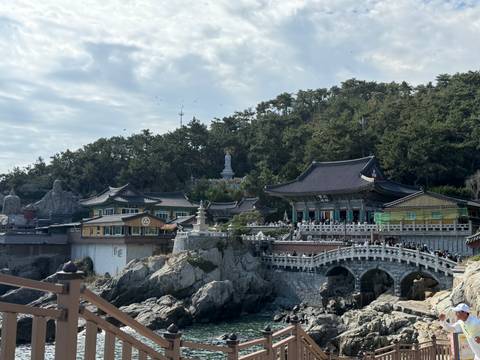       Coastal Buddhist temple complex built on rocky cliffs with many visitors crossing a stone bridge.
  