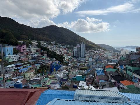       Colourful hillside neighbourhood of Gamcheon Culture Village beneath a lush mountain and blue sky.
  