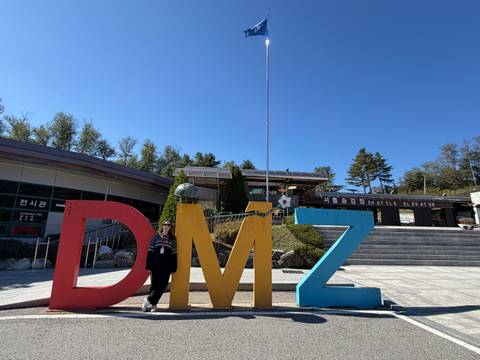       Tourist poses beside large colourful DMZ letters under a clear blue sky.
  