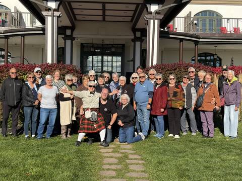       Large tour group poses smiling on a lawn in front of a historic building on a sunny day.
  