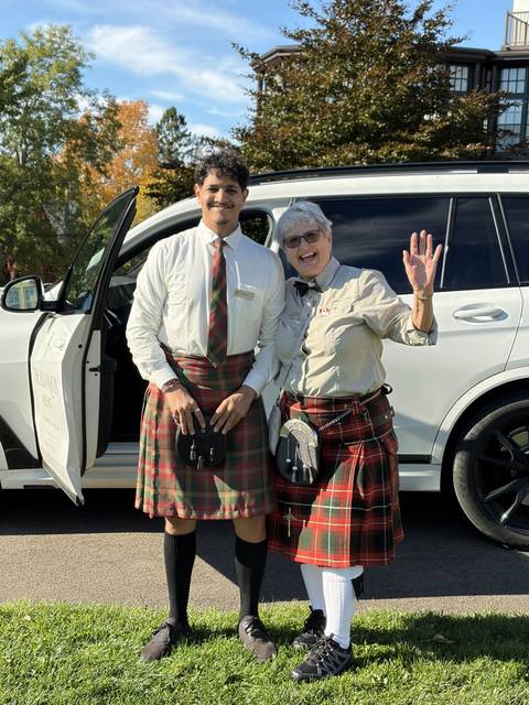       Two guides in kilts stand smiling and waving beside a white SUV.
  