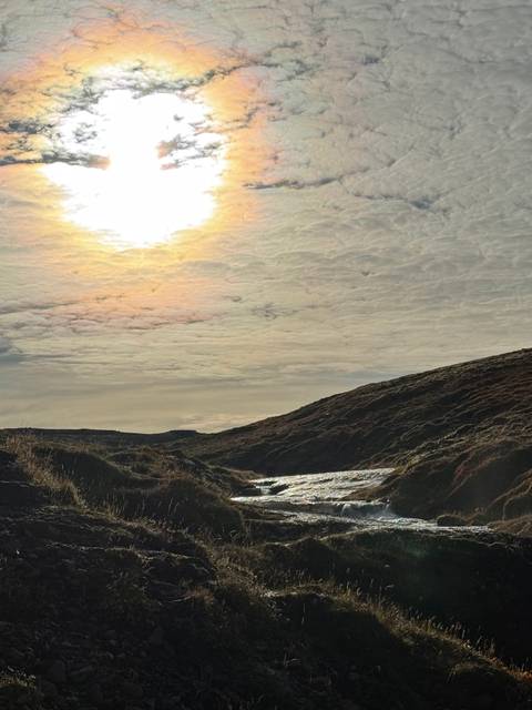       Soft sunrise light filters through dramatic cloud cover above a rugged grassy hillside and small stream.
  