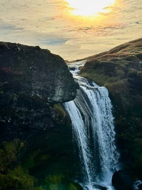       A narrow waterfall cascades between dark volcanic cliffs into a rocky pool at dusk.
  