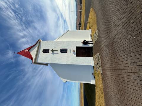       A small white church with a red roof stands isolated under a wide blue sky as a visitor pauses by the door.
  