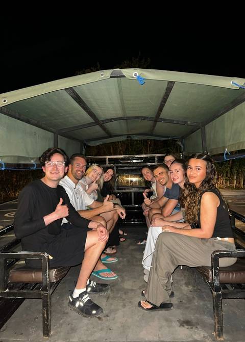       Group of young travelers smiling and giving thumbs-up while seated in the back of an open truck at night.
  