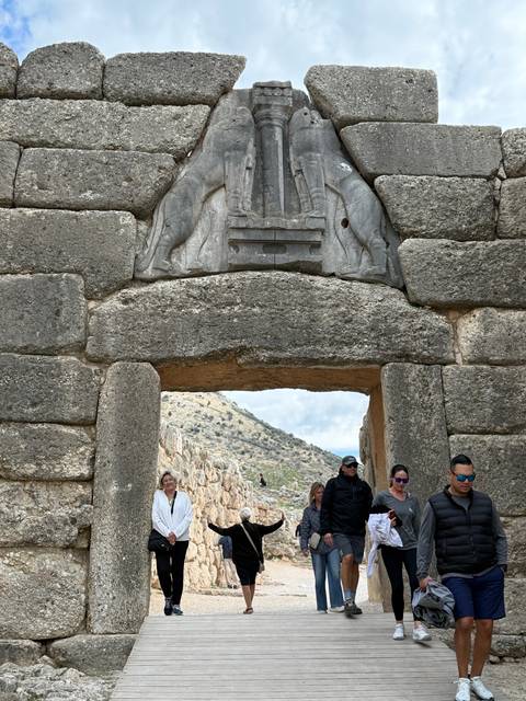       Visitors stand beneath the Lion Gate entrance of ancient Mycenae with carved stone lions above.
  