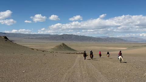       Riders on horseback traverse vast open steppe toward distant Altai mountains under scattered clouds.
  