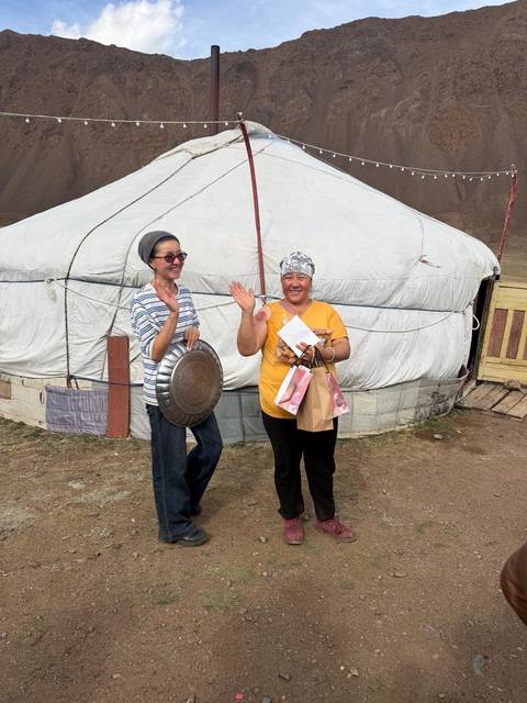       Two smiling Mongolian women wave outside a white felt ger, holding gifts after a visit.
  