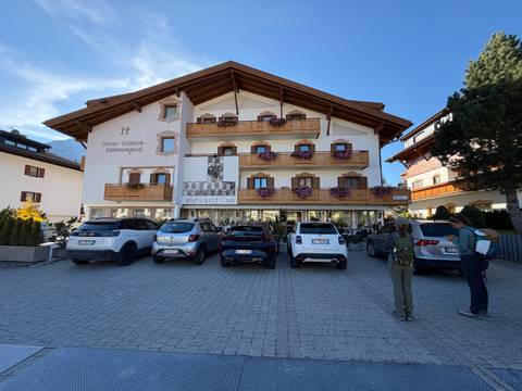       Alpine hotel with flower-decked balconies and parked cars, hikers preparing in front.
  