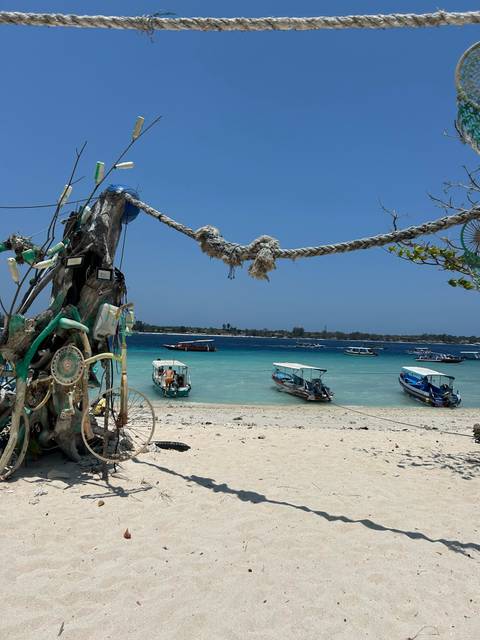       Tropical beach scene with turquoise water, several small motorboats anchored near shore and a driftwood structure decorated with ropes and debris.
  