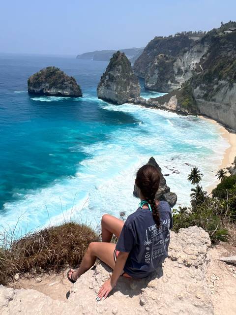       A traveller sits on a cliff edge overlooking brilliant blue waves and a secluded white-sand beach bordered by high limestone cliffs.
  