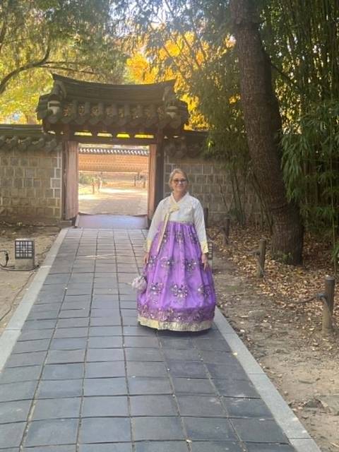       Woman wearing a purple hanbok stands in a traditional Korean courtyard surrounded by stone walls and bamboo.
  