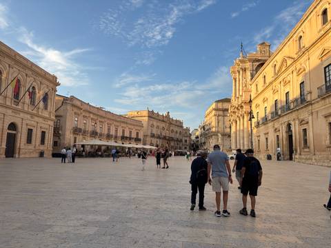       Evening light bathes elegant baroque buildings around a spacious stone piazza bustling with pedestrians.
  