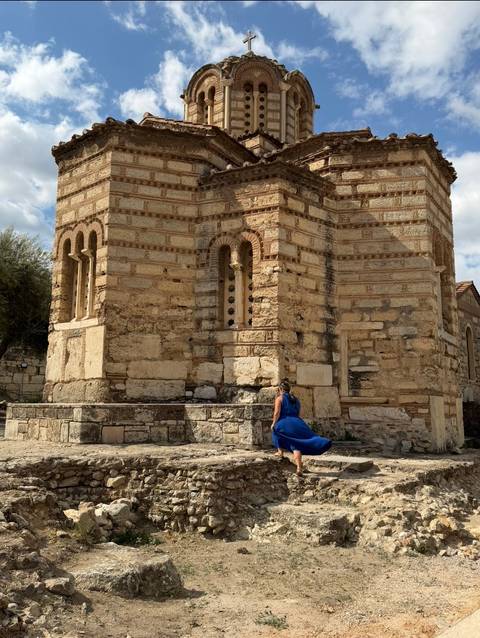       Woman in flowing blue dress exploring an ancient stone church with arched windows and weathered masonry.
  