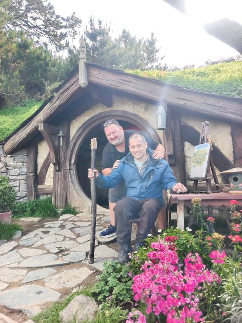       Two friends pose outside a hobbit-style round door in lush green surroundings.
  
