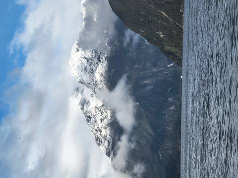       Snow-dusted peaks rise above misty fjord waters under patchy clouds.
  