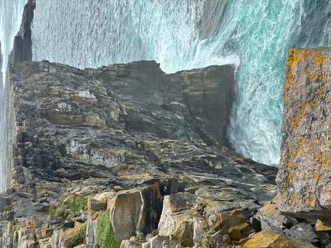       A dramatic sheer sea cliff rises above turquoise waves crashing at its base.
  