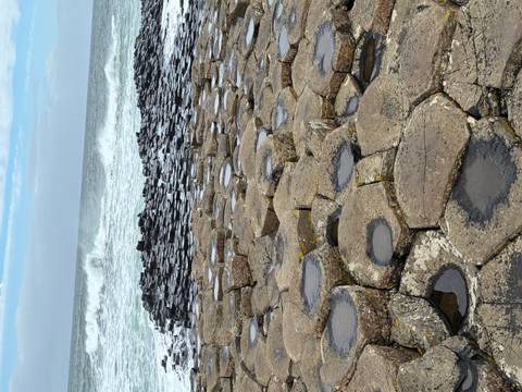       Hexagonal basalt columns of a coastal rock formation stretch toward the sea with waves crashing beyond.
  