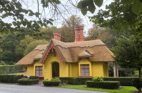       A quaint bright-yellow thatched cottage with ornate gables sits amid lush gardens and trees.
  