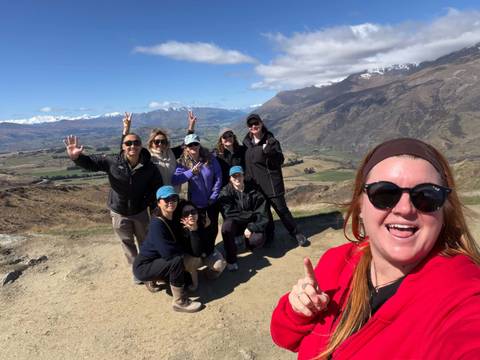       Smiling group of travelers pose at a scenic mountain viewpoint with snow-capped peaks and a broad valley behind them under blue skies.
  