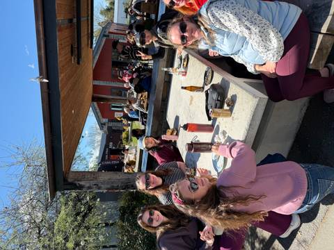       Outdoor communal table where travelers enjoy food and drinks in the sun, laughing and chatting at a casual eatery.
  