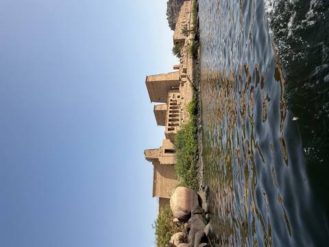       Ancient sandstone temple structures on an island, viewed from the water with gentle ripples under a clear blue sky
  