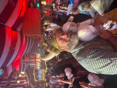       Man in Bavarian costume wearing horned helmet cuddles a plush sheep at a lively Oktoberfest beer hall.
  