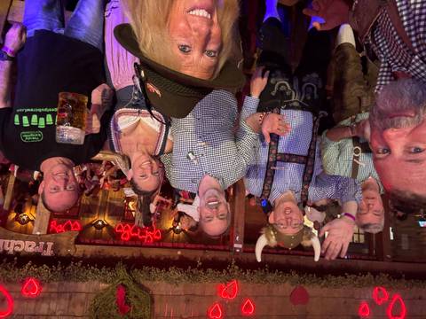       Smiling group in traditional Bavarian attire raise hands and beer steins inside a decorated beer tent.
  