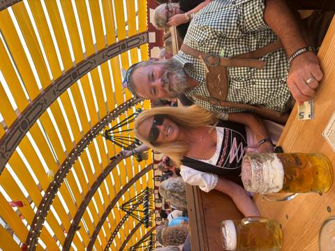       Couple in dirndl and lederhosen sit at a wooden table with a giant beer mug inside a brightly striped beer tent.
  