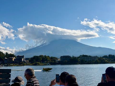       Mount Fuji capped by dramatic lenticular cloud looms above lakeside town and speeding yellow boat.
  