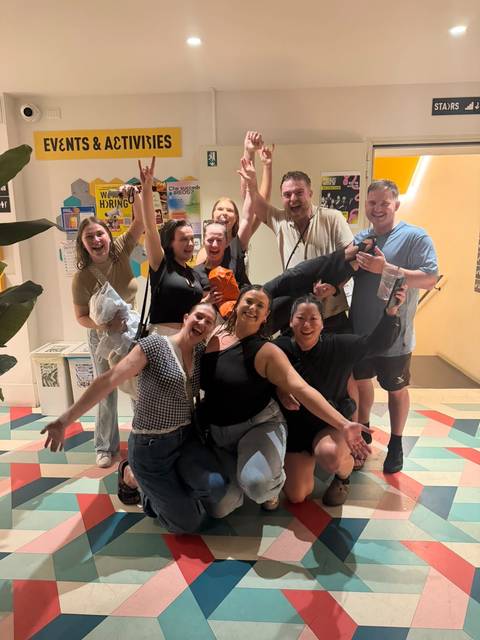       Friends celebrate indoors, laughing and posing energetically in a brightly lit hallway.
  