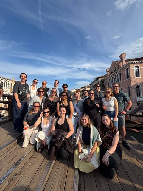       Large travel group poses on a Venetian wooden bridge with historic pastel buildings behind them.
  