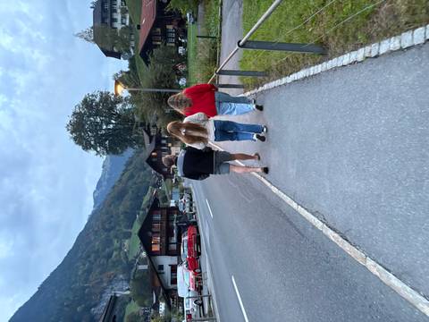       Three friends walk along a quiet alpine village road at dusk with mountains in the distance.
  