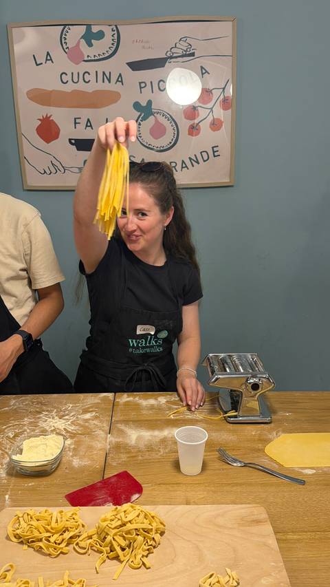       A smiling woman in an apron lifts fresh pasta sheets beside a metal pasta machine during a cooking class.
  