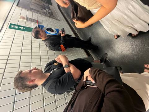       Travellers wait on a tiled underground platform corridor beside signage and emergency phone.
  