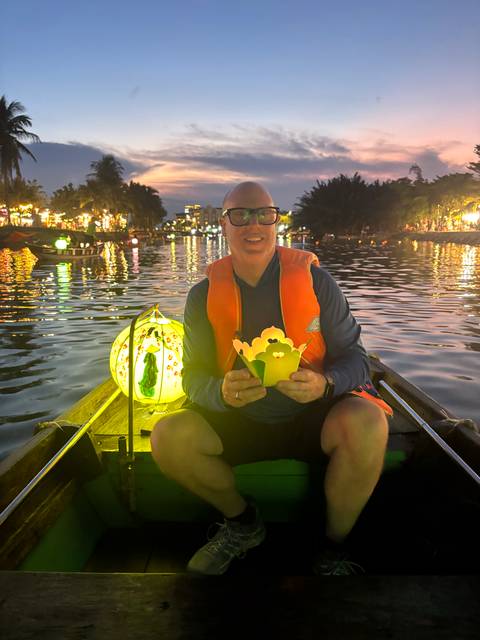       Man in a small boat at dusk holding a paper lantern on a river lit by colourful lights
  