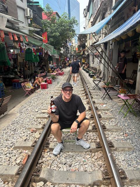       Tourists relaxing on fold-out chairs along Hanoi Train Street while a man poses with a beer on the rails
  