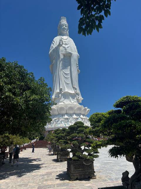       Base of the towering white Lady Buddha statue framed by lush greenery under clear sky
  