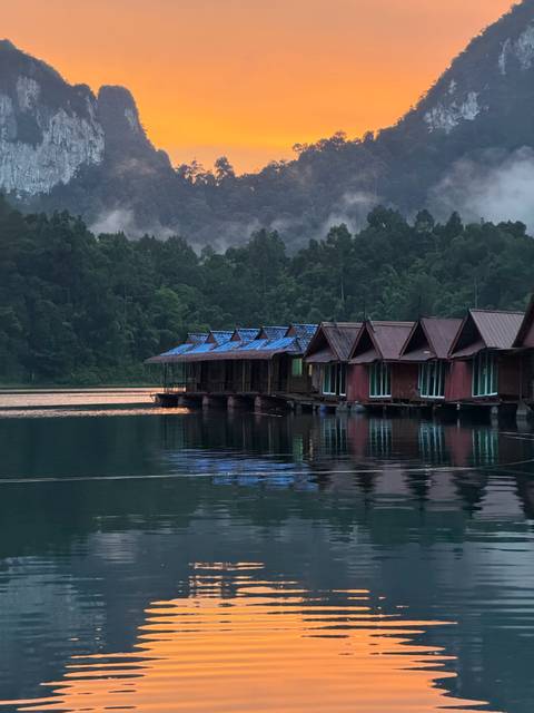       Row of rustic floating wooden bungalows reflected on a calm emerald lake at dusk with forested hills beyond.
  