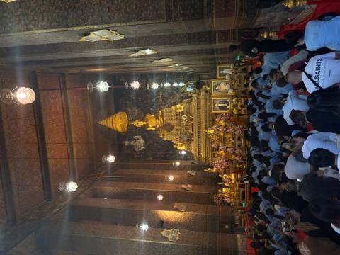       Interior of a grand Thai temple with a towering golden Buddha statue as worshippers sit on the floor in prayer.
  