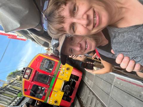       Selfie of travellers inches from a colorful train passing through a narrow market street.
  