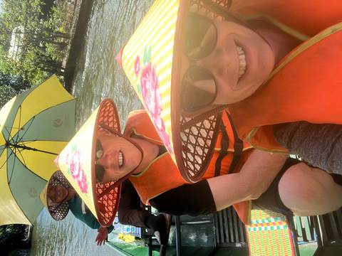       Travellers in bright life vests and conical hats paddling down a shaded river under umbrellas.
  