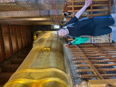       Visitor posing beside the enormous golden Reclining Buddha inside a richly decorated hall.
  